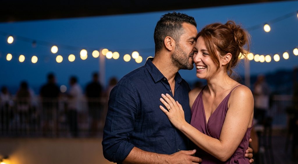 Couple d’amants complices qui rient sur un rooftop guirlandé au-dessus de la mer, le soir