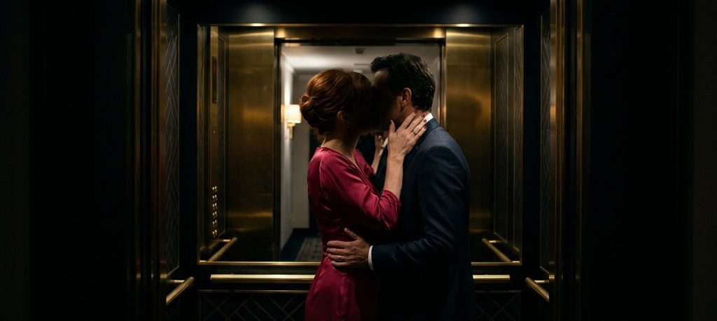 Amants enlacés qui s’embrassent dans l’ascenseur d’un hôtel, elle en robe framboise