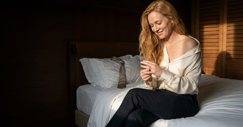 Femme mariée assise sur un lit d’hôtel, chemise de soie ouverte sur l’épaule, qui fait tourner son alliance en souriant