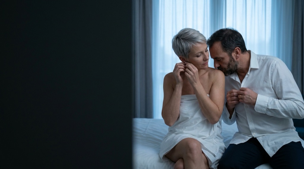 Amants dans une chambre d’hôtel au petit matin, il l’embrasse sur l’épaule avant de partir