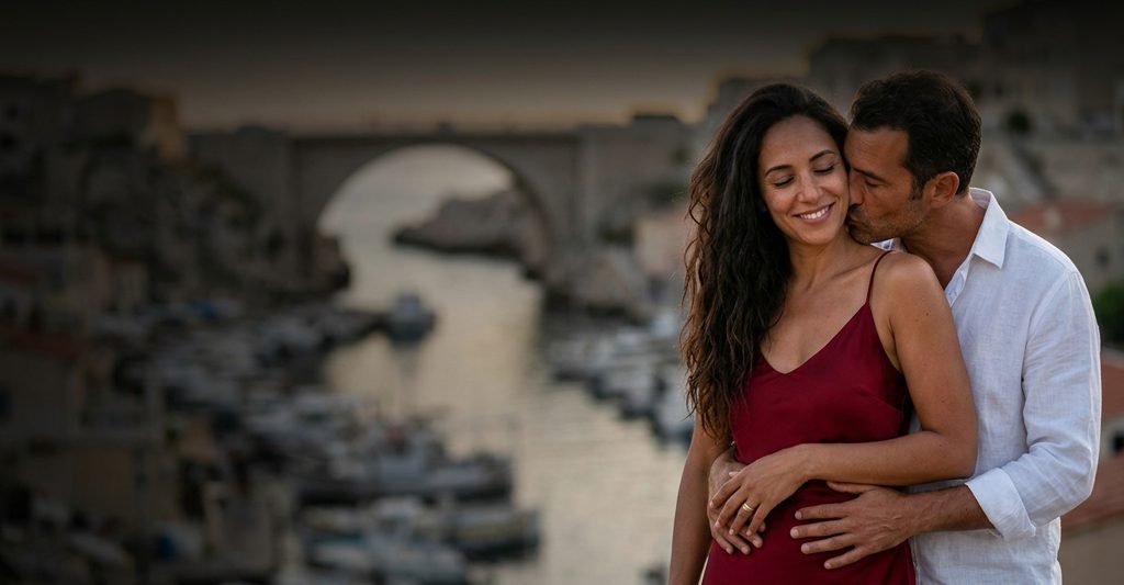 Couple d’amants enlacés au bord d’un petit port de Marseille, barques en contrebas, elle en robe rouge